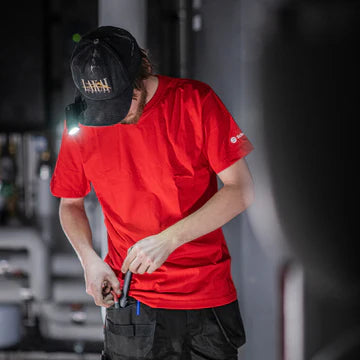 A man wearing a red shirt and a black cap with an LED clip light attached to the brim is working in an industrial setting. He is looking down and using a tool, with the LED light providing bright, focused illumination for his task. The environment includes various pipes and equipment, indicating a technical or maintenance-related activity.