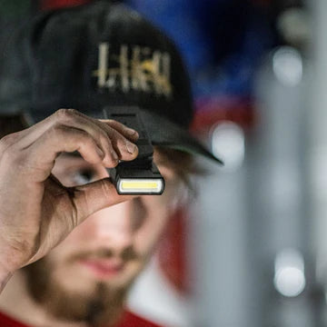 A man wearing a black cap is holding a compact black LED clip light close to his face, showcasing the light's design and LED strip. The background is slightly blurred, emphasizing the focus on the man and the light. The man appears to be in an industrial or workshop setting.