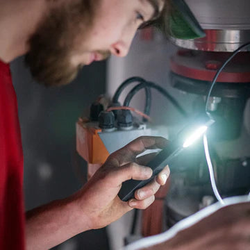 A man in a red shirt uses a black LED penlight to illuminate a piece of industrial equipment. He is focused on inspecting or repairing the machinery, with the penlight providing a bright, concentrated beam of light to aid his work. The environment suggests a technical or maintenance setting.