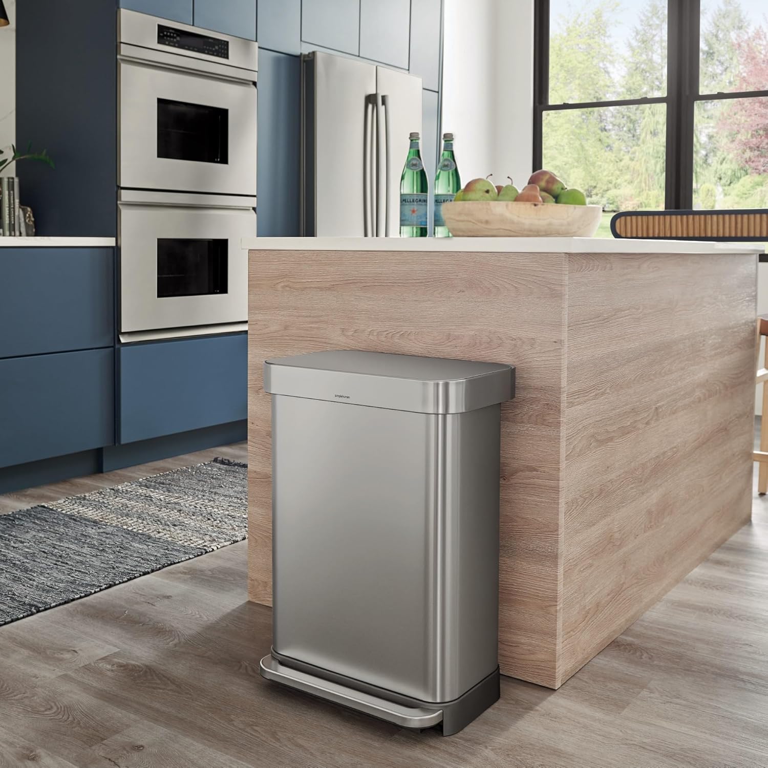 Stainless steel rectangular trash can with a foot pedal, placed in a modern kitchen next to a wooden island. The sleek design blends seamlessly with the contemporary decor, featuring blue cabinetry, stainless steel appliances, and a bright, open space.