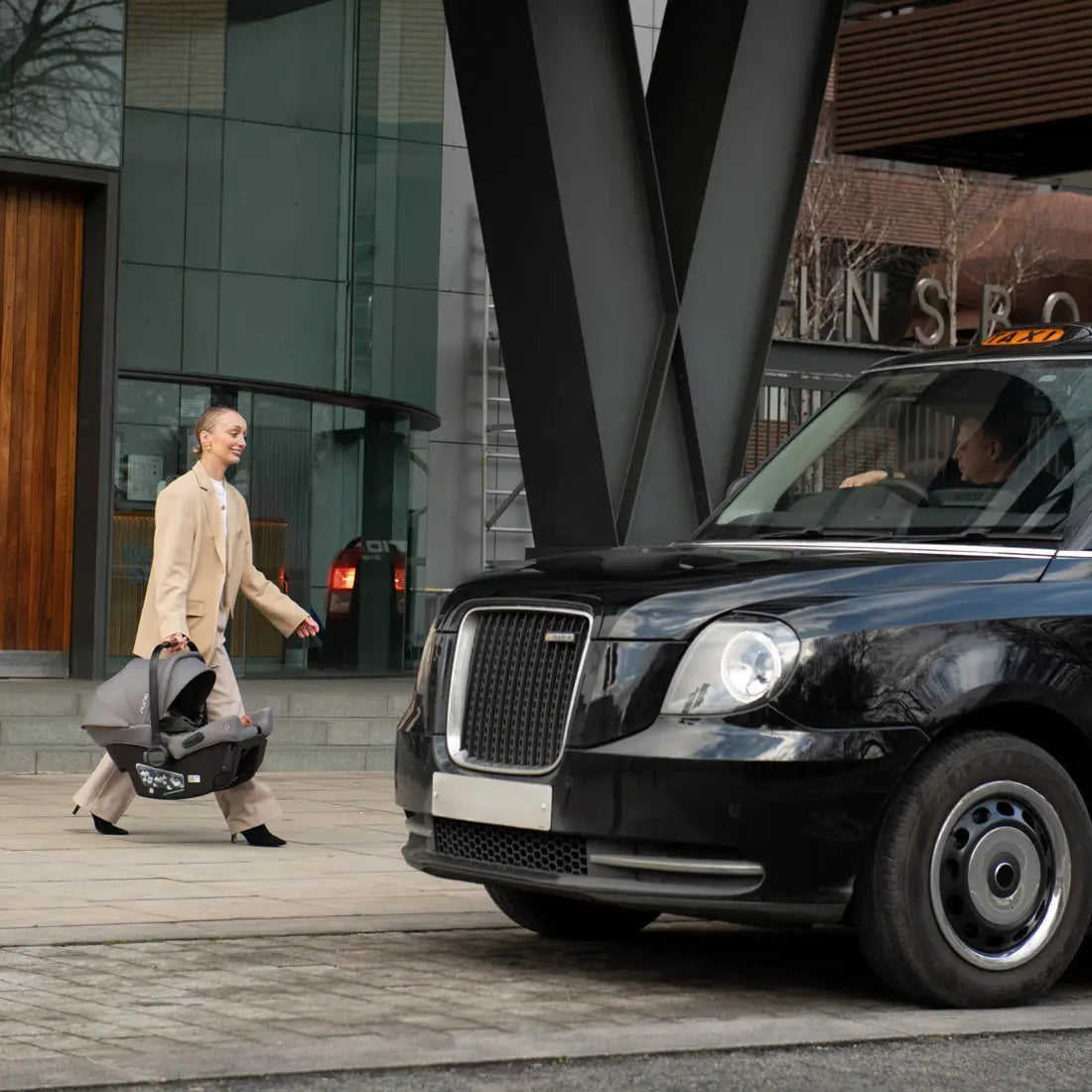 A woman walks toward a black taxi while carrying a baby car seat, set in front of a modern building with glass panels and a wooden door.