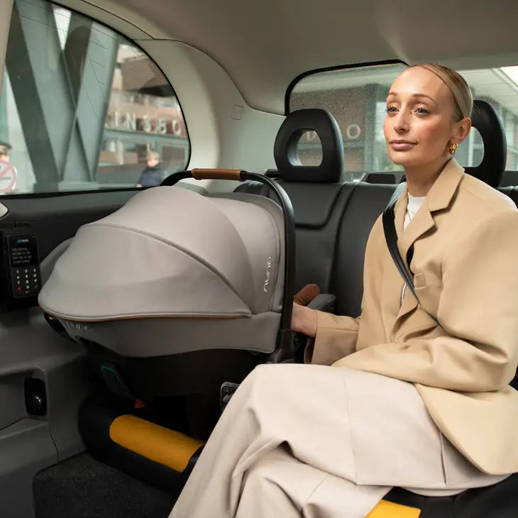 A woman in a beige outfit sits in the back seat of a vehicle, next to a securely installed Nuna infant car seat with a grey canopy and brown handle, showcasing a travel-friendly car seat setup.