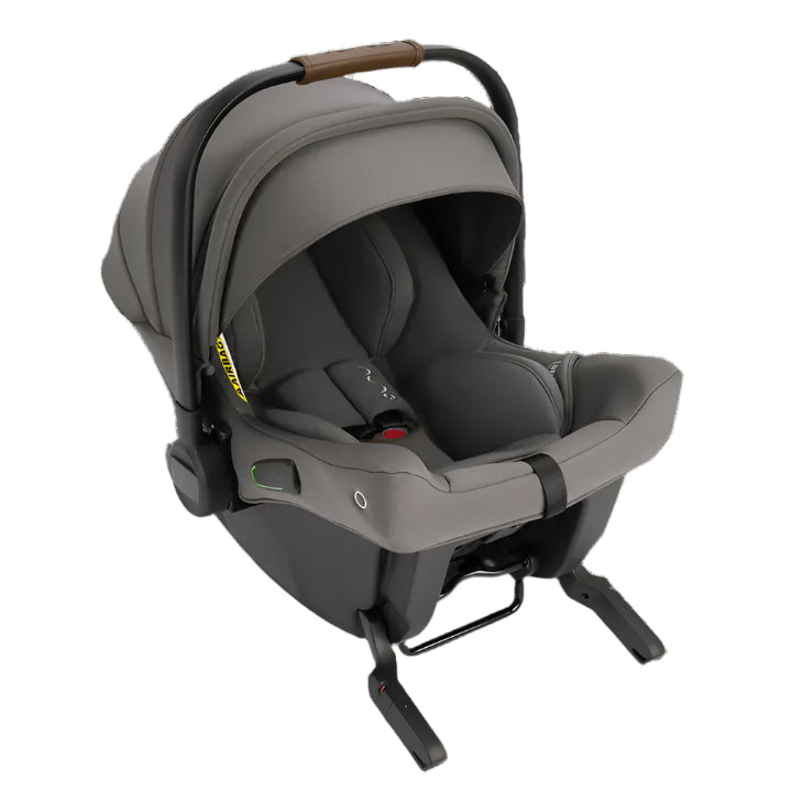 A Nuna infant car seat in grey is shown with an extended canopy, brown leatherette handle, visible green safety indicators, and fold-out stability legs for secure vehicle installation.