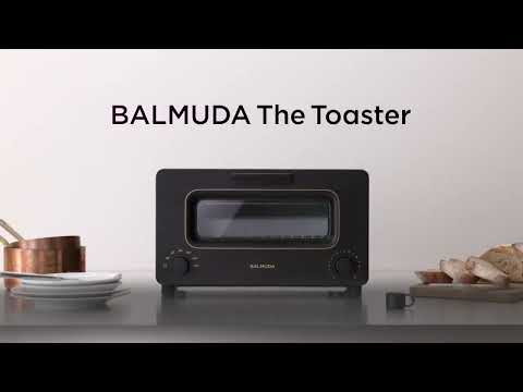 The image is a screenshot from a YouTube video titled "BALMUDA The Toaster." The video features a Balmuda toaster placed on a kitchen countertop, with the product prominently displayed in the center of the frame. The toaster is black with gold accents and has a sleek, modern design.