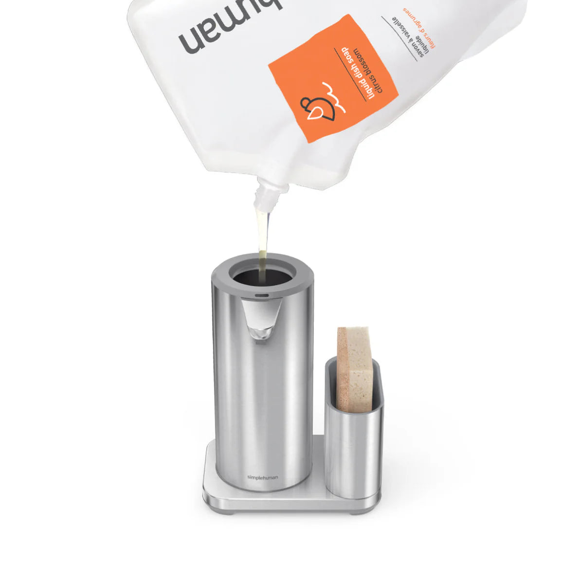 A stainless steel automatic soap dispenser being refilled with liquid soap from a large refill pack. The dispenser is cylindrical and positioned on a rectangular base, with a sponge holder and sponge beside it. The refill pack is white with an orange label, and soap is being poured into the top of the dispenser.