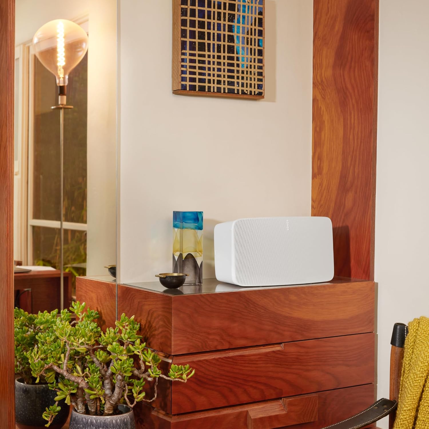 A Sonos wireless speaker in white placed on a wooden sideboard in a stylishly decorated room. The setting features a modern aesthetic with a large mirror, decorative items such as a colorful glass vase, and a green potted plant adding warmth to the space. The speaker seamlessly blends into the decor, complementing the wooden textures and ambient lighting. The scene highlights the speaker's compact and elegant design, making it a perfect fit for contemporary interiors.