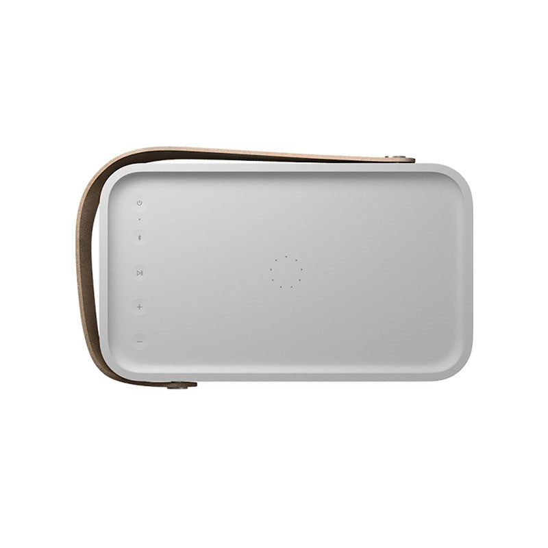 This image shows the **top view of a Bang & Olufsen (B&O) portable speaker**. The speaker has a **minimalist and clean design** with a light grey surface. The **control buttons** for volume adjustment, play/pause, and Bluetooth pairing are neatly arranged along the left edge. A central circular pattern likely indicates the speaker’s **multi-function touch control area**. The **leather strap** attached to the side of the speaker adds to the portability and aesthetic appeal. The overall design emphasizes simp