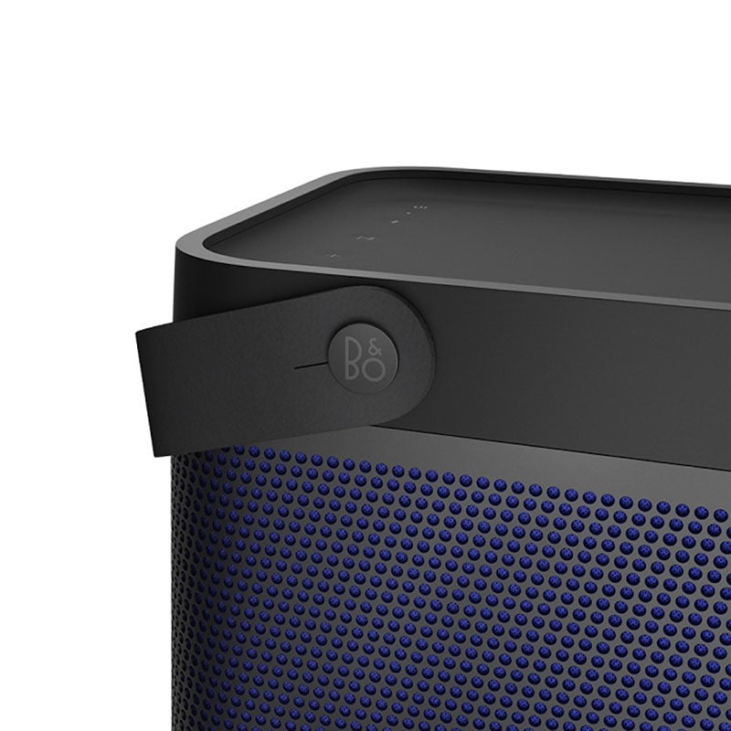 This close-up image shows a detailed view of the **Bang & Olufsen (B&O) portable speaker**, specifically focusing on the **strap** with the **B&O logo** embossed on it. The **perforated grille** is clearly visible, featuring a **blue color** under the black exterior frame, adding a distinct, premium look. The top of the speaker has a smooth surface with subtle **control indicators**, reflecting the minimalist design approach typical of B&O products. The combination of materials and design elements suggests