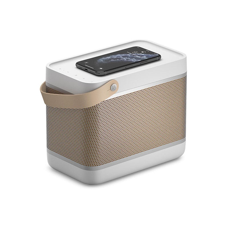 This image features a **Bang & Olufsen (B&O)** portable speaker in a **light grey and beige variant**. The speaker has a **minimalist design**, with a **light perforated grille** in a beige color. On top of the speaker, there is a **smartphone** placed, indicating **wireless charging capability**. The speaker is equipped with a **leather strap** on the side, which provides portability and a touch of luxury, and the iconic **B&O logo** is embedded on the strap. This model exemplifies the brand’s emphasis on