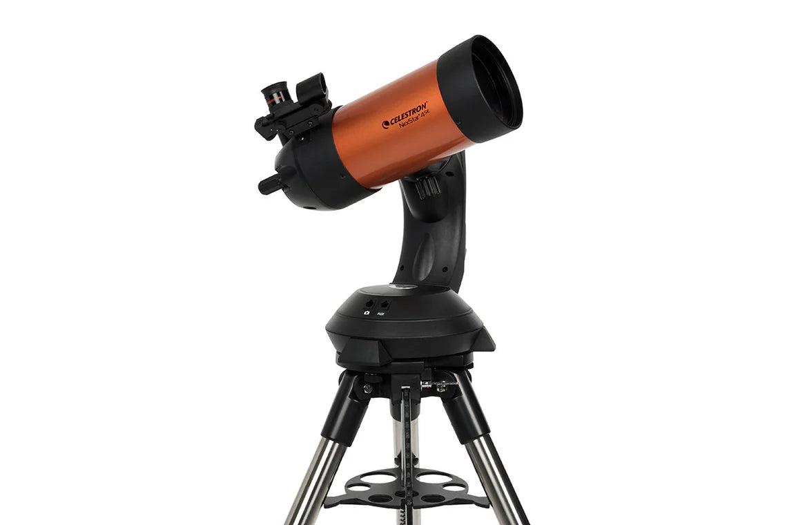 A Celestron NexStar 4SE telescope with an orange cylindrical tube mounted on a black computerized base and a silver tripod. The telescope is angled slightly upward for observation. The eyepiece and a small finderscope are attached to the back of the telescope tube. The base provides advanced tracking and alignment capabilities