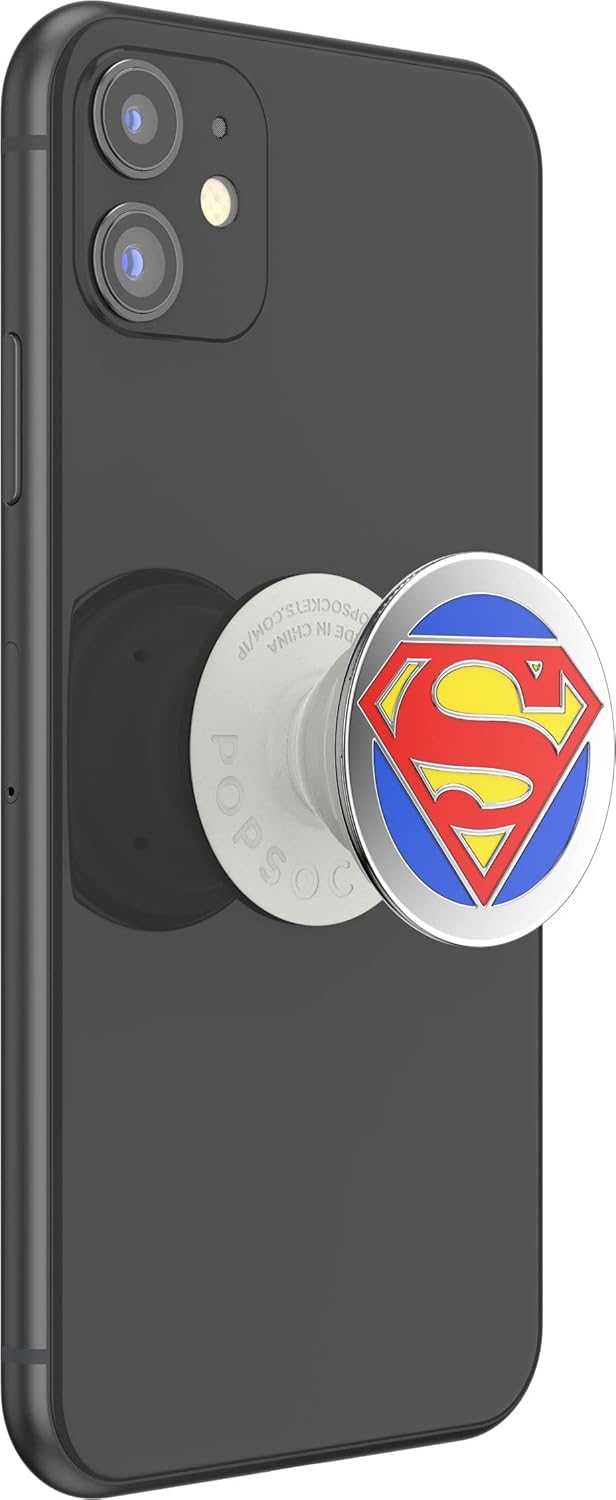 A black smartphone with a PopSockets Enamel Swappable Grip featuring the Superman logo attached to the back, shown in an extended state. The grip displays the iconic Superman "S" shield in red and yellow on a blue background, encased in a shiny metal border. The extended position provides a functional grip for holding and supporting the smartphone, adding both style and convenience with its swappable and expandable features.