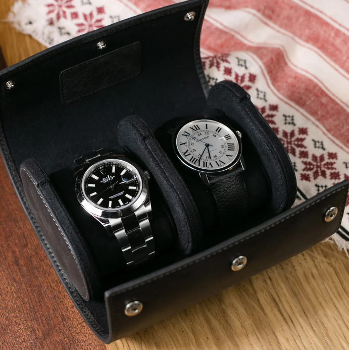 Two elegant wristwatches secured in a black leather travel case, with one watch featuring a black dial and metal band and the other showcasing a white dial with Roman numerals and a black leather strap, placed on a wooden surface beside a patterned fabric.