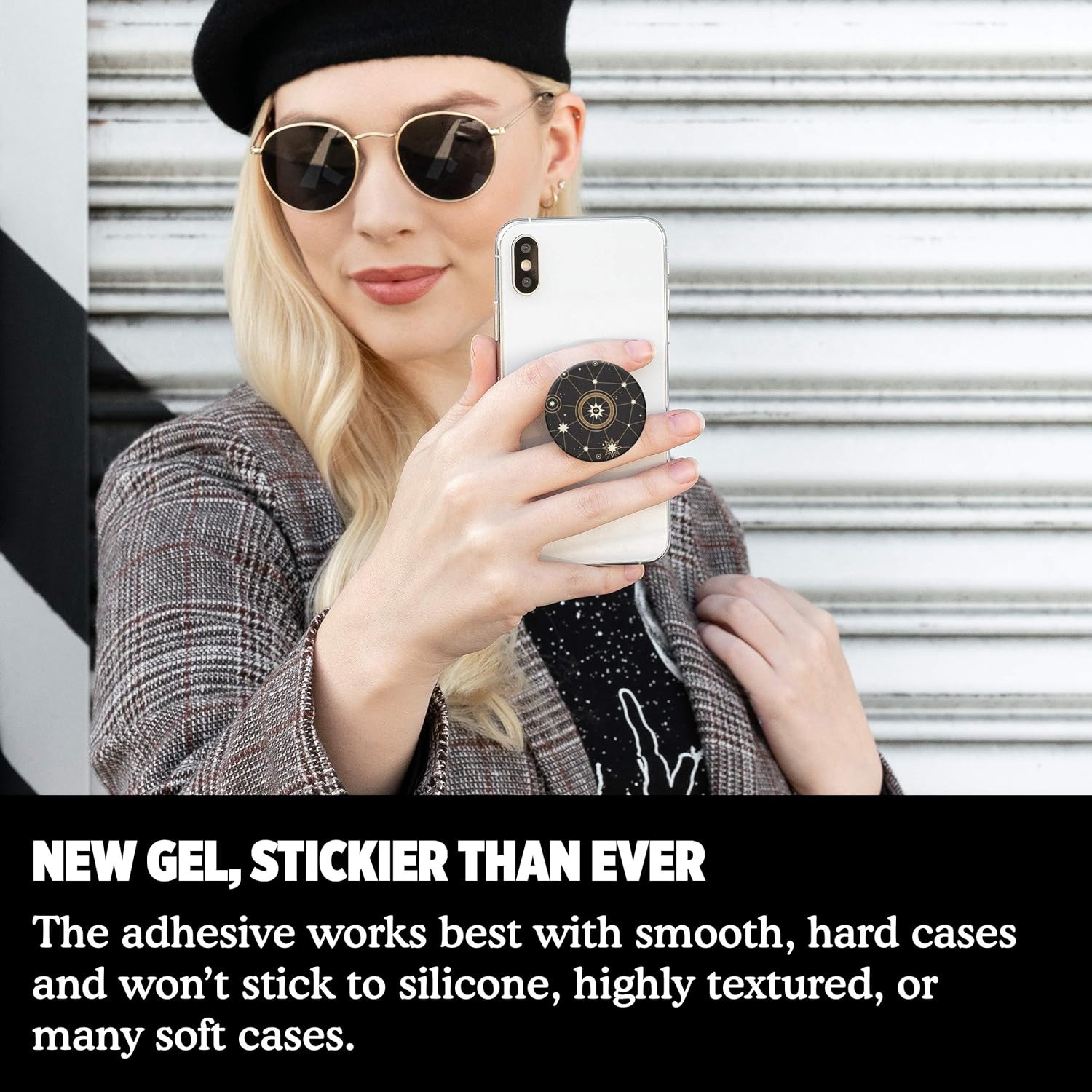 A person wearing a black beret and round sunglasses is holding a white phone with a PopSockets grip featuring a star chart design, taking a selfie. The person is dressed in a plaid blazer.