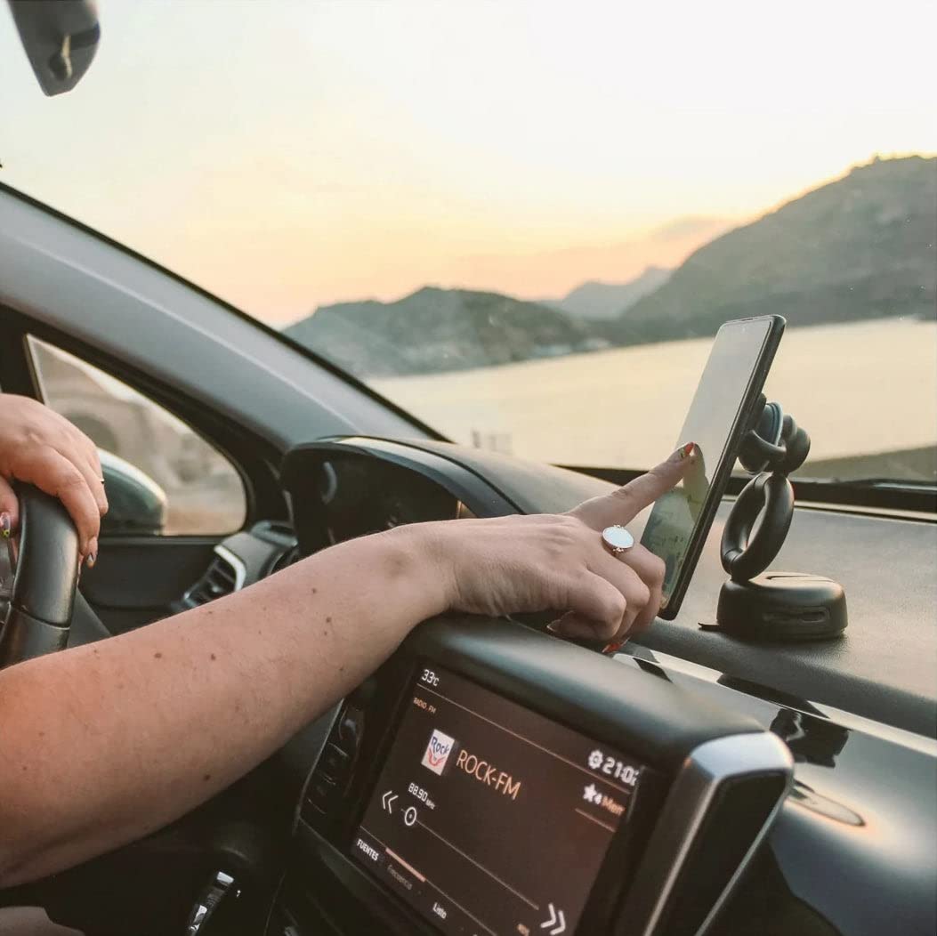 A person using a smartphone mounted on a PopSockets Multi-Use G1 Dash Mount attached to a car dashboard. The mount securely holds the phone in an upright position, allowing for easy access and visibility while driving. The person's hand is interacting with the phone screen, demonstrating the mount's functionality and stability. The background shows a scenic view of a lake and hills during sunset.