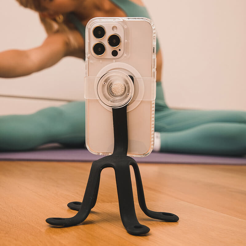 A smartphone with a clear case mounted on a black PopSockets Flex Mount, positioned on a wooden floor. The Flex Mount has a tripod-like design with three flexible legs providing stability. In the background, a person in workout attire is seen stretching on a yoga mat, indicating the use of the mount for hands-free viewing or recording during exercise. The setup highlights the mount's versatility and practicality for various activities.