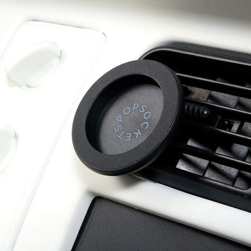 A Black MagSafe Car Vent PopMount 2 is shown attached to a car's air vent. The mount features a circular design with the PopSockets logo embossed on the inner surface. The black finish of the mount matches the air vent, creating a sleek and integrated look. The mount is securely attached to the vent, providing a stable platform for holding a MagSafe-compatible device for hands-free use while driving.