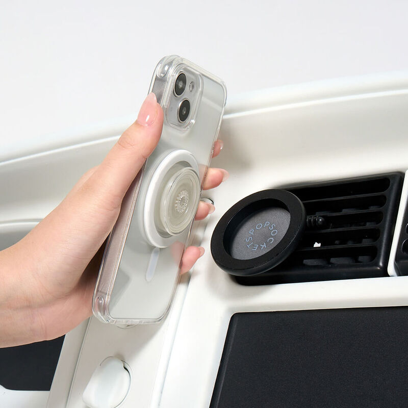 A hand is shown holding a smartphone with a clear case, which has a clear PopSocket grip attached to its back. The hand is positioning the phone near a Black MagSafe Car Vent PopMount 2 that is securely attached to a car's air vent. The mount features a circular design with the PopSockets logo embossed on the inner surface. The setup demonstrates the ease of attaching the phone to the car vent mount for hands-free use while driving.