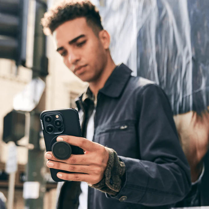 A man is shown outdoors, holding a smartphone with a black case. The phone has a PopGrip MagSafe in black attached to its back. The man is wearing a dark jacket and is looking at his phone, demonstrating the functionality and sleek design of the PopGrip while using his phone. The urban background suggests he is in a city setting.