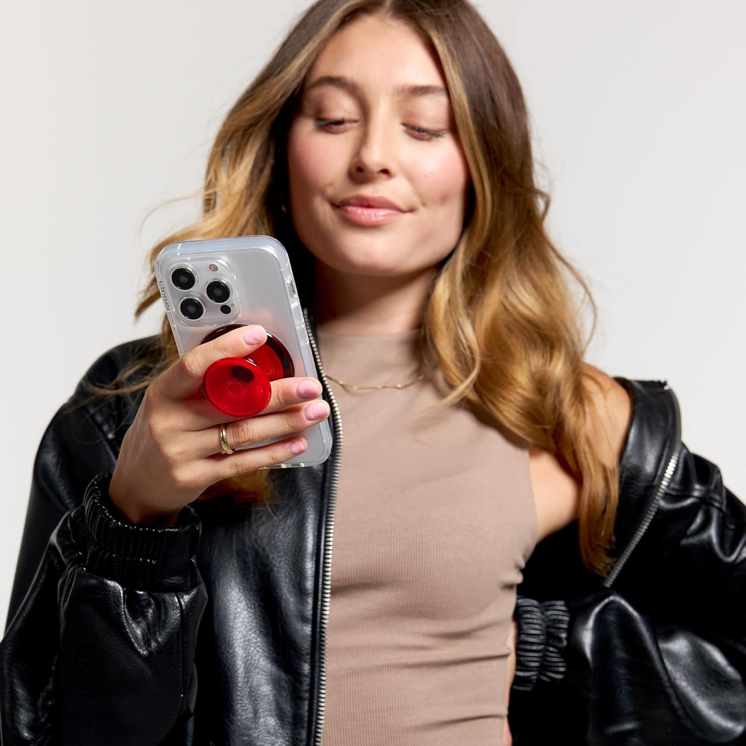 A woman with long, wavy hair holds a smartphone with a clear case and a red PopSocket grip. She is dressed in a black leather jacket and beige top, looking at her phone with a relaxed expression.