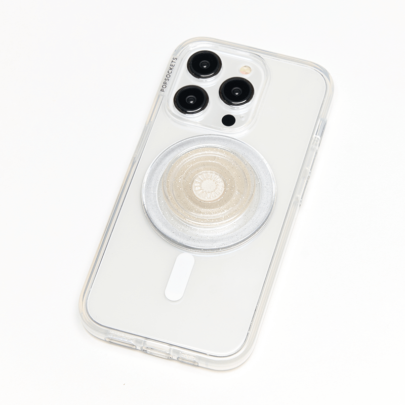 A smartphone with a clear case is shown lying flat with a PopSockets Clear Glitter PopGrip for MagSafe attached to its back. The PopGrip features a round, flat surface with a clear, glittery finish, positioned centrally on the phone's back. The phone's camera lenses are visible at the top, and the sparkling design of the PopGrip complements the phone's aesthetics while providing a functional grip.