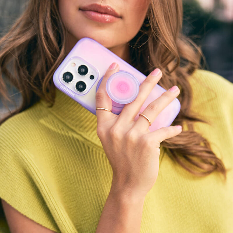 A woman with wavy hair is holding a smartphone with a gradient pink and purple case. The phone has an Aura PopGrip for MagSafe attached to its back, featuring a gradient color pattern transitioning from pink in the center to purple on the edges. The woman is showcasing the PopGrip with her fingers holding it, highlighting its functionality and stylish design. She is wearing a yellow sleeveless top, and her fingernails are manicured in a natural color.