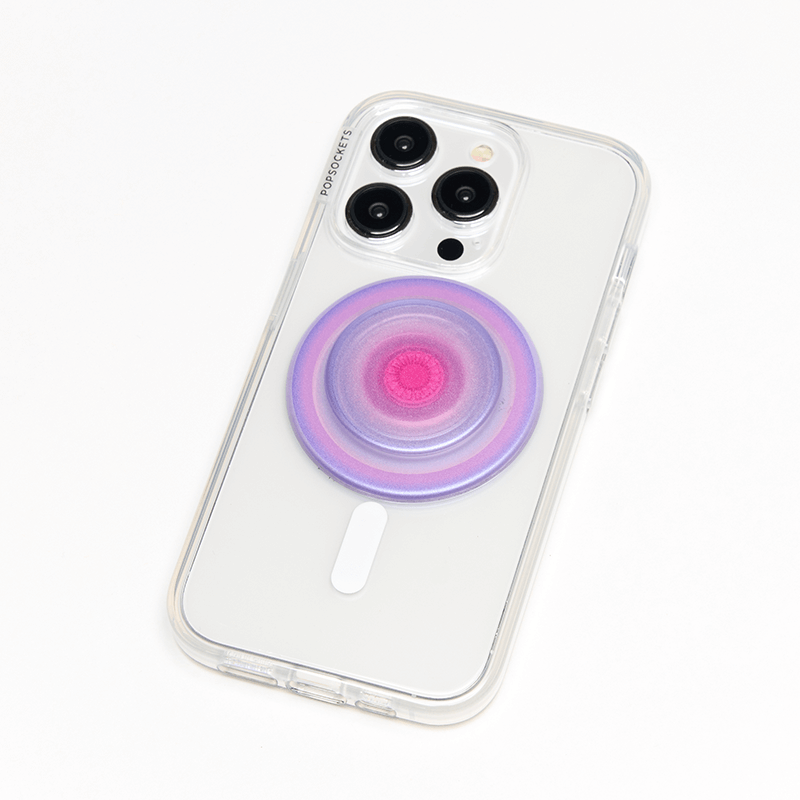 A smartphone with a clear case is shown lying flat with an Aura PopGrip for MagSafe attached to its back. The PopGrip features a round, flat surface with a gradient color pattern transitioning from pink in the center to purple on the edges. The phone's camera lenses are visible at the top, and the colorful design of the PopGrip complements the phone's aesthetics while providing a functional grip.