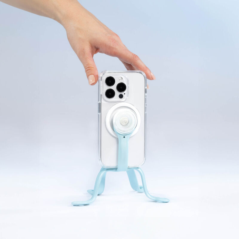  A hand is adjusting a smartphone held by a PopSockets Flex Mount in Blue Sigh color. The smartphone is in a clear case with a white PopGrip attached to the back. The mount's flexible legs are positioned to support the phone upright on a flat surface. The setup is displayed against a light blue background.