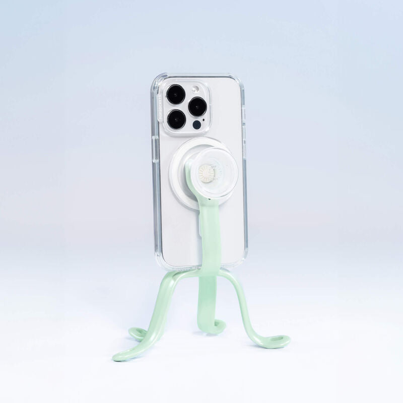  PopSockets Flex Mount in Honey Dew color holding a smartphone in a clear case with a white PopGrip attached to the back. The mount's flexible legs are positioned to support the phone upright on a flat surface. The phone is shown from the back, highlighting the PopGrip and the mint-green colored flexible mount's design. The setup is displayed against a light blue background.