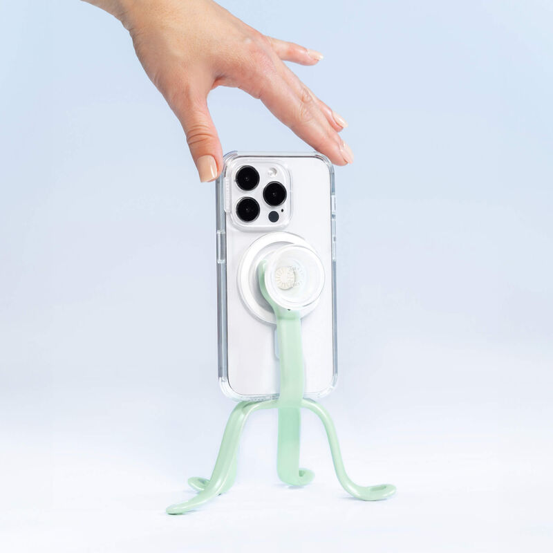  A hand is adjusting a smartphone held by a PopSockets Flex Mount in Honey Dew color. The smartphone is in a clear case with a white PopGrip attached to the back. The mount's flexible legs are positioned to support the phone upright on a flat surface. The setup is displayed against a light blue background.