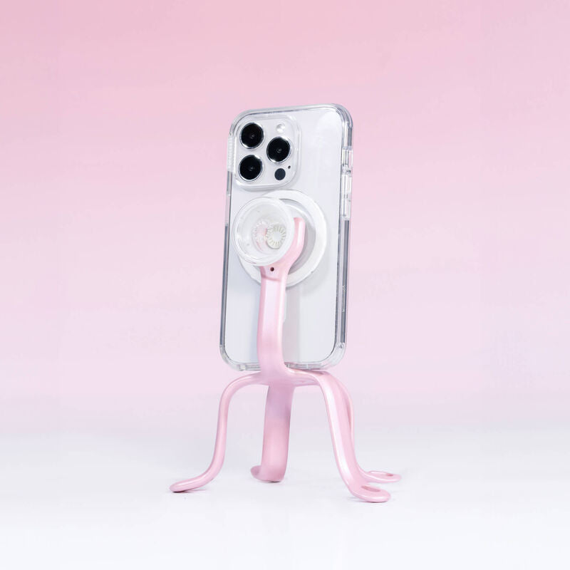  PopSockets Flex Mount in Pinky color holding a smartphone in a clear case with a white PopGrip attached to the back. The mount's flexible legs are positioned to support the phone upright on a flat surface. The phone is shown from the back, highlighting the PopGrip and the light pink colored flexible mount's design. The setup is displayed against a light pink background.