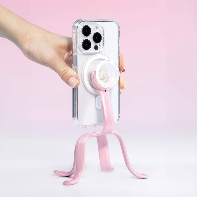 A hand is adjusting a smartphone held by a PopSockets Flex Mount in Pinky color. The smartphone is in a clear case with a white PopGrip attached to the back. The mount's flexible legs are positioned to support the phone upright on a flat surface. The setup is displayed against a light pink background