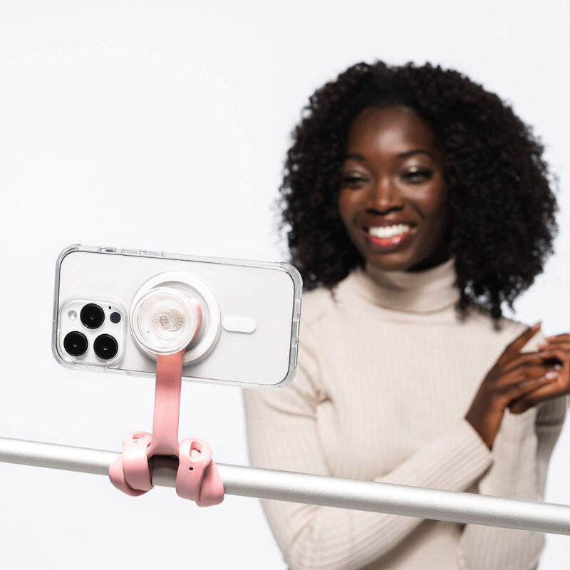  PopSockets Flex Mount in Pinky color holding a smartphone in a clear case with a white PopGrip attached to the back. The mount's flexible legs are wrapped around a horizontal metal bar, securing the smartphone in a horizontal position. In the background, a woman with curly hair wearing a light-colored turtleneck sweater is smiling and looking towards the camera. The setup demonstrates the mount's ability to hold the smartphone securely for video calls or recording.