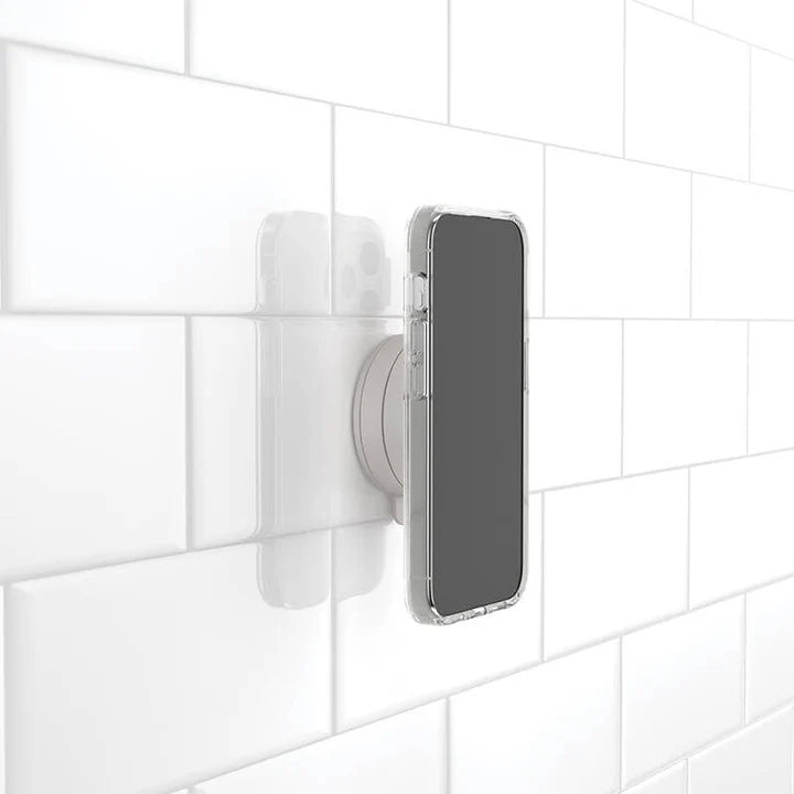 A smartphone is attached to a white tiled wall using a PopSockets Suction Mount for MagSafe in the color black. The phone is in a clear case, and the mount holds it securely against the smooth, tiled surface, highlighting the mount's strong suction and compatibility with MagSafe technology