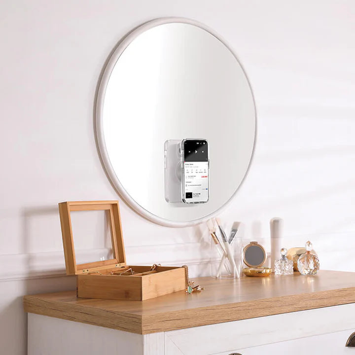 A round mirror is mounted on a wall above a wooden dresser. Attached to the mirror is a smartphone held by a black PopSockets Suction Mount for MagSafe. The dresser has various items on it, including a wooden jewelry box, makeup brushes, a small tray with perfume bottles, and other decorative objects. The black mount securely holds the phone on the mirror's surface, showcasing its functionality and design.