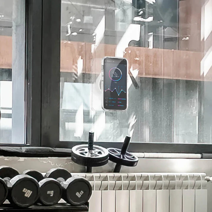 A smartphone is attached to a window using a black PopSockets Suction Mount for MagSafe. The phone displays a fitness tracking app with various graphs and data. Below the window, there are gym equipment items, including dumbbells and ab rollers, indicating that the setting is a gym. The mount securely holds the phone on the window, demonstrating its strong suction capability and suitability for use in a workout environment.