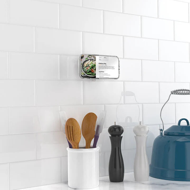 A smartphone is attached to a white tiled kitchen wall using a black PopSockets Suction Mount for MagSafe. The phone displays a recipe for chicken pho. Below the phone, there is a container holding wooden cooking utensils, and nearby are a pepper grinder, a salt shaker, and a utensil holder. The setup illustrates the mount's practicality for hands-free use in a kitchen setting, making it easy to follow recipes while cooking.