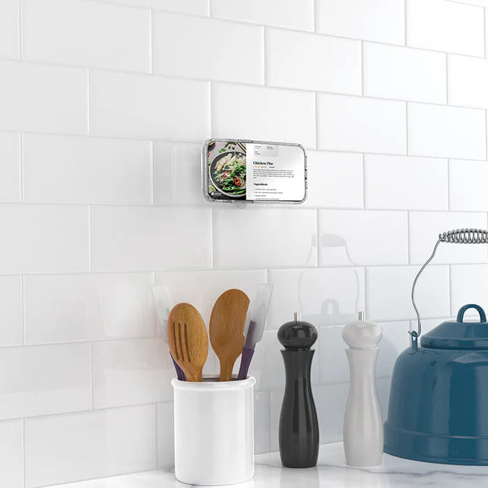 A smartphone is attached to a white tiled kitchen wall using a dusty rose PopSockets Suction Mount for MagSafe. The phone displays a recipe for chicken pho. Below the phone, there is a container holding wooden cooking utensils, and nearby are a pepper grinder, a salt shaker, and a blue teapot, illustrating the mount's practical use for hands-free cooking in a kitchen setting.