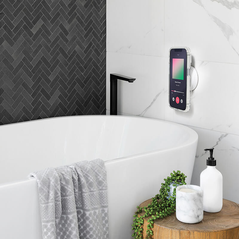 PopSockets Suction Mount for MagSafe in Horchata color attached to a tiled wall in a bathroom. The mount is holding a smartphone next to a bathtub, demonstrating its secure attachment and practical use in a bathroom setting. The background features a mix of dark and light tiles, with a sleek faucet, a towel draped over the bathtub, and various bathroom accessories nearby.