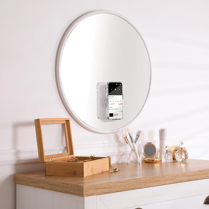  A round mirror is mounted on a wall above a wooden dresser. Attached to the mirror is a smartphone held by a PopSockets Suction Mount for MagSafe in the color horchata. The dresser has various items on it, including a wooden jewelry box, makeup brushes, a small tray with perfume bottles, and other decorative objects. The PopSockets mount securely holds the phone on the mirror's surface, showcasing its functionality and design.