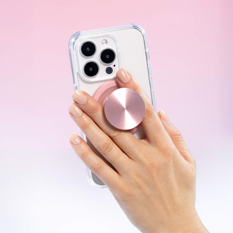 A hand is shown holding a smartphone with a clear case, which has a PopSockets MagSafe PopGrip in dusty rose color attached to its back. The PopGrip is extended, allowing the fingers to grip it securely. The glossy, slightly reflective soft pink surface of the PopGrip complements the phone's aesthetics. The phone's camera lenses are visible at the top, and the hand's nails are manicured in a light, natural color.