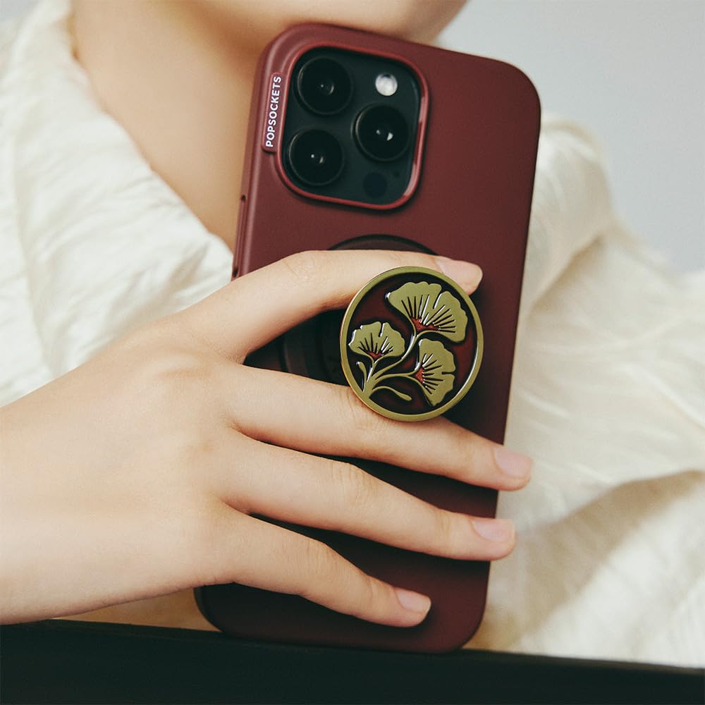 This image features a **dark red phone case** with a **PopSocket** attached to the back, showcasing an elegant design of **green ginkgo leaves with a red branch**. The PopSocket adds both style and functionality, providing a comfortable grip for holding the phone securely. The user's hand is elegantly holding the phone, with the PopSocket in use.