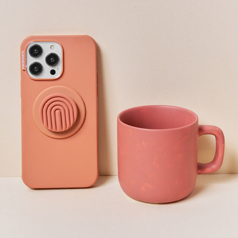 A terracotta-colored phone case with a PopOut Terracotta Curves PopGrip attached is displayed next to a matching terracotta-colored mug. The PopGrip features a terracotta-colored design with curved lines, complementing the minimalist and stylish aesthetic of both the phone case and the mug