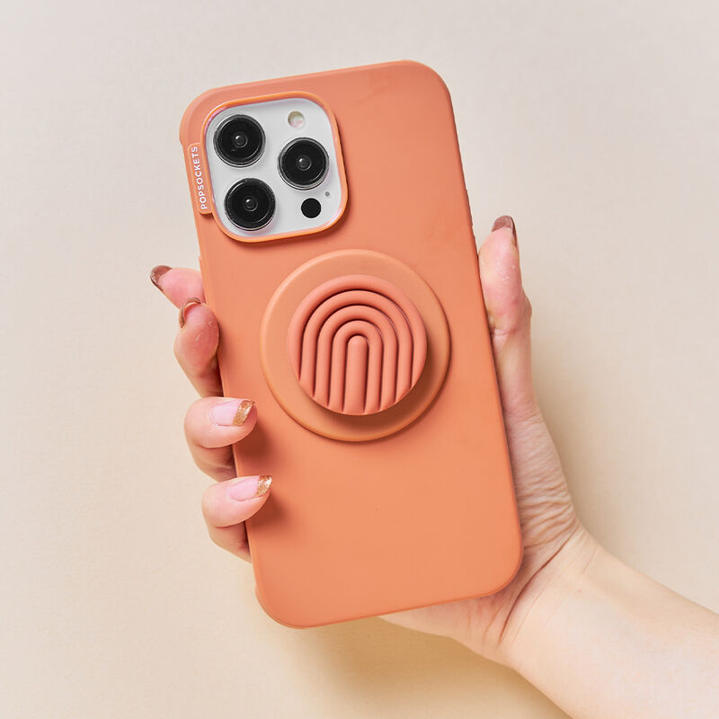 A hand with a manicure is holding a smartphone with a terracotta-colored case and a PopOut Terracotta Curves PopGrip attached. The PopGrip features a terracotta-colored design with curved lines, providing a comfortable and stylish grip for the device