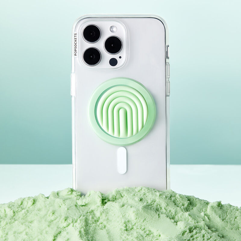 A clear phone case with a PopOut Curves Matcha Dew PopGrip for MagSafe is displayed standing upright in a pile of light green powder. The PopGrip features a light green color with curved lines, complementing the pastel aesthetic of the scene