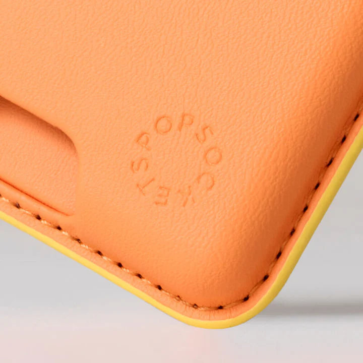 A close-up image of the Softgoods PopWallet for MagSafe in cantaloupe color. The texture of the material is visible, along with the embossed "PopSockets" logo near the bottom corner. The stitching around the edges is clearly shown, highlighting the wallet's detailed craftsmanship. The bright cantaloupe color is complemented by a yellow lining along the edges.