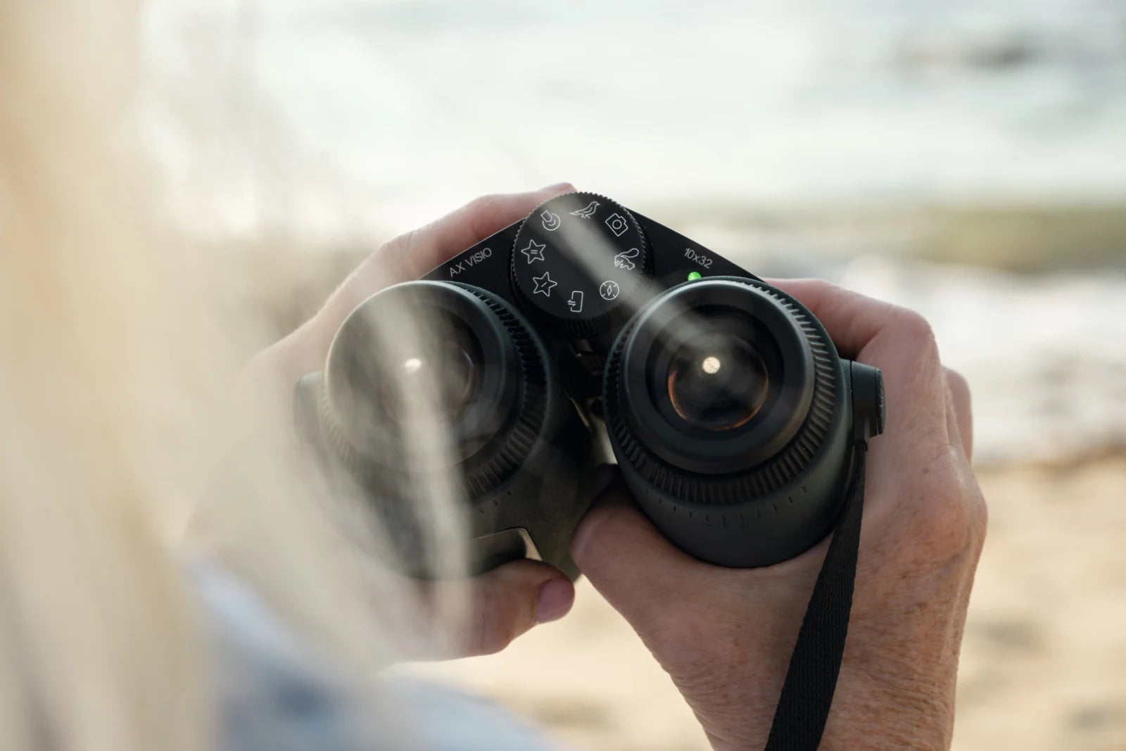 Person holding AX Visio binoculars with SWAROVISION technology, looking at a distant view near the beach.