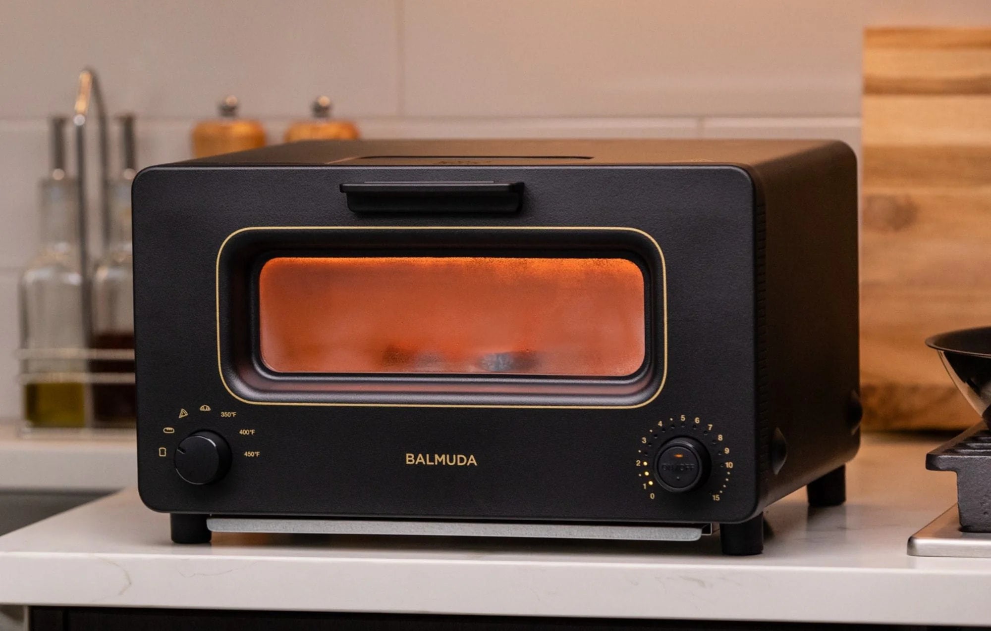 : The image shows a Balmuda toaster in a matte black finish with gold accents, placed on a kitchen countertop. The toaster's rectangular viewing window is illuminated with a warm orange glow from the heating elements inside, indicating that it is in use. The brand name "BALMUDA" is centered below the window in gold lettering.