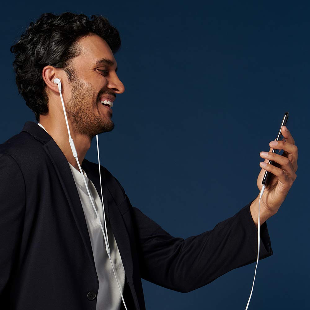 The image shows a man smiling and using wired earphones connected to his smartphone. He is engaging in what appears to be a video call or conversation while holding his phone. The backdrop is a solid dark blue, and the man is dressed casually in a blazer, giving a polished yet relaxed appearance.