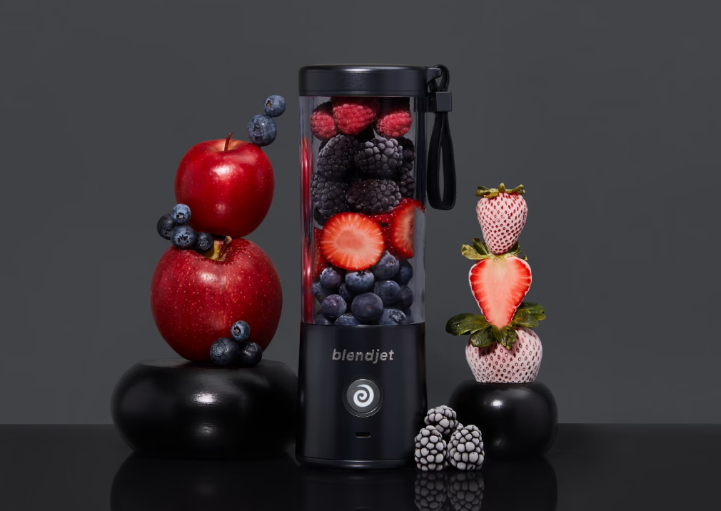 A sleek black portable blender filled with fresh berries, including strawberries, blackberries, blueberries, and raspberries, placed on a glossy black surface. On the left, two stacked red apples decorated with blueberries sit on a black base. On the right, skewered white strawberries and halved red strawberries are stacked on another black base. The background is a dark gray, complementing the vibrant fruit colors. The blender features a circular power button with a swirl design and a carrying strap attach