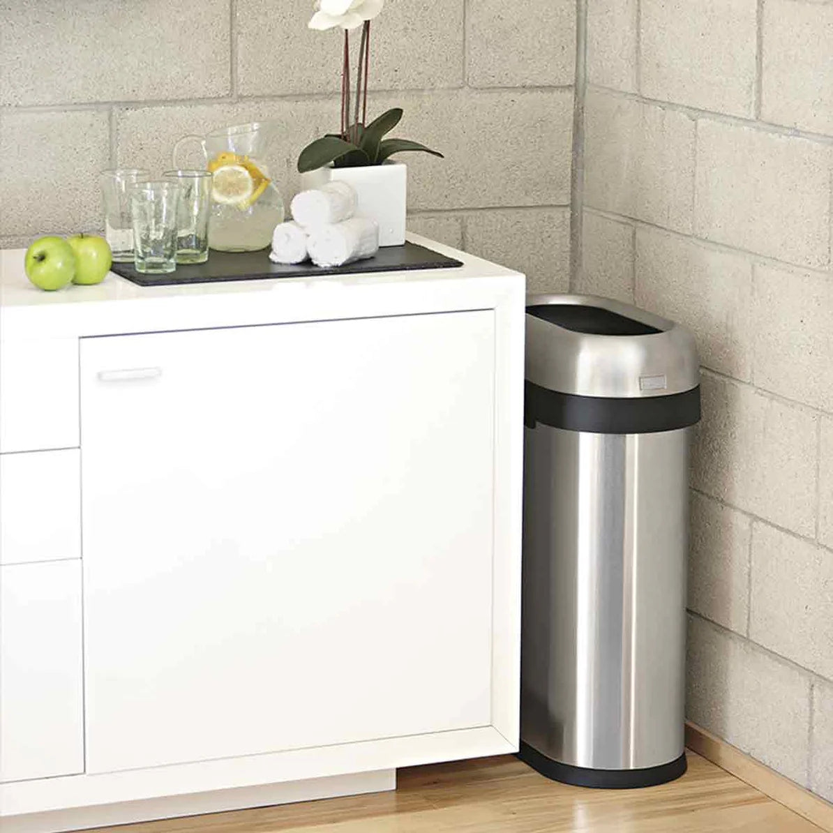 The image shows a stainless steel trash can with a black, oval-shaped open-top lid, positioned neatly in the corner of a modern, minimalist kitchen or utility area. The trash can is placed next to a white cabinet with a wooden countertop, which holds a few decorative items, including a plant, glassware, and apples. The setting features a concrete block wall and light wood flooring, creating a clean and contemporary environment. The trash can's sleek design fits well with the stylish decor, offering both fun